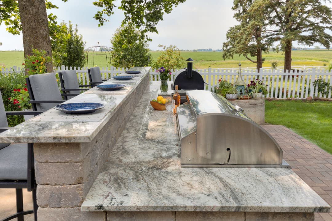 Outdoor kitchen with granite countertop, modern grill, and seating area in serene garden setting.