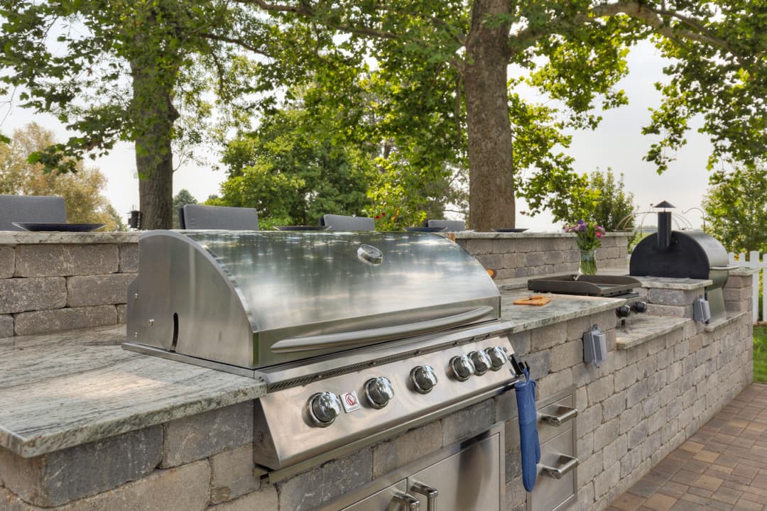 Outdoor kitchen with a stainless steel grill, stone countertops, and shaded seating area.